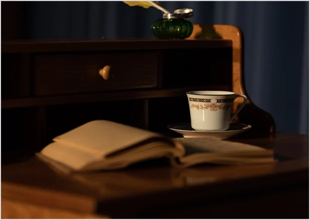 An open vintage book on a dark wooden desk next to a classic teacup and an inkwell with a quill, representing historical manuscript preservation and traditional publishing heritage.