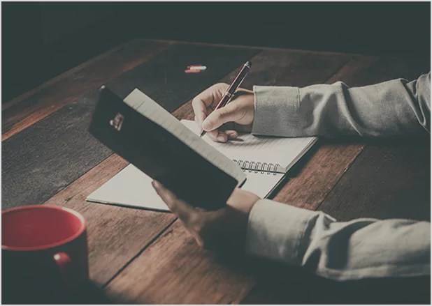 A person holding a pencil while checking a printed manuscript against a digital tablet, representing professional editorial verification and cross-platform publishing quality control.