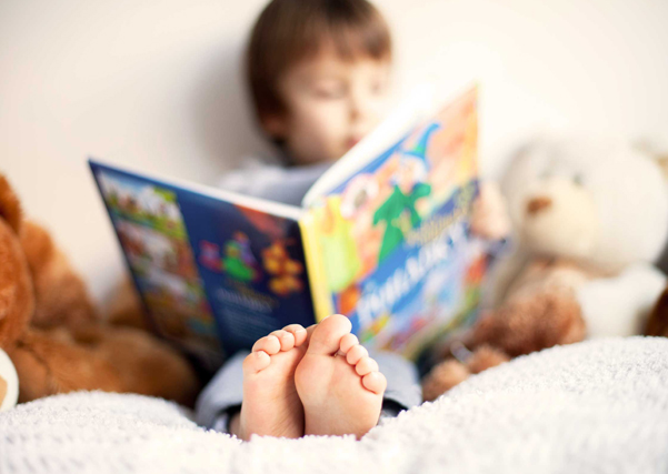 A young child reading a colorful picture book in bed with teddy bears in the background.
