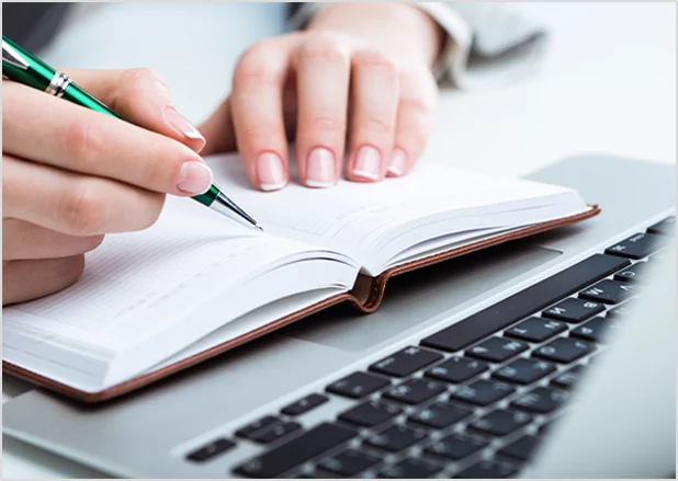 Close-up of a person’s hands using a green and silver pen to write in a brown leather-bound planner resting on a silver laptop.
