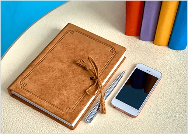 A brown leather-bound journal with a tassel next to a smartphone and pen, representing integrated digital and traditional publishing solutions.