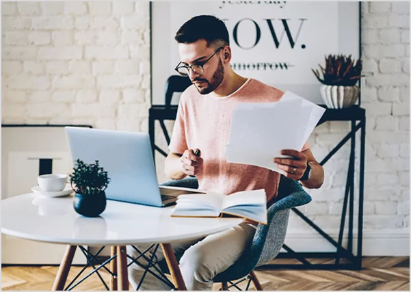 A man with glasses sitting at a white table, holding a manuscript and reviewing his laptop, representing professional manuscript formatting and digital publishing preparation services.