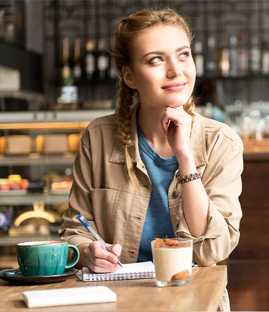 A thoughtful woman with braided hair writing in a notebook at a cafe, highlighting personalized book coaching services.
