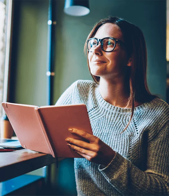 A young woman with glasses and a grey sweater looking up thoughtfully while holding a small peach-colored notebook in a brightly lit cafe.