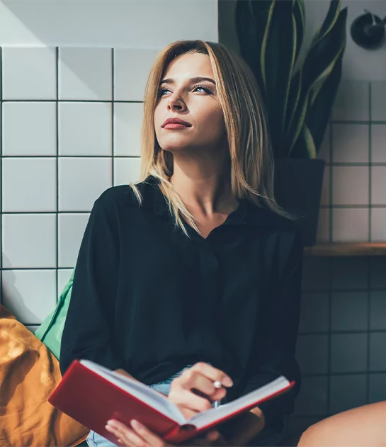 A thoughtful blonde woman in a black shirt holding a red notebook while looking out a window, representing creative professional development and author mindset coaching.