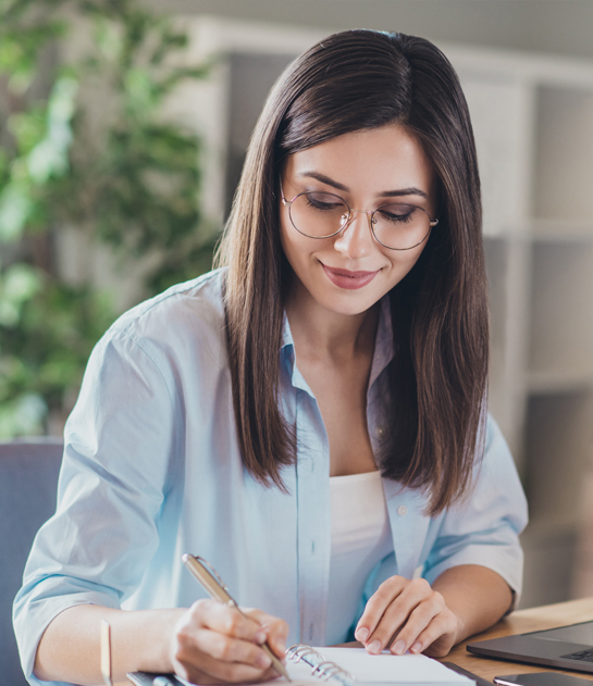 A young woman with glasses and brown hair writing in a notebook at a desk.