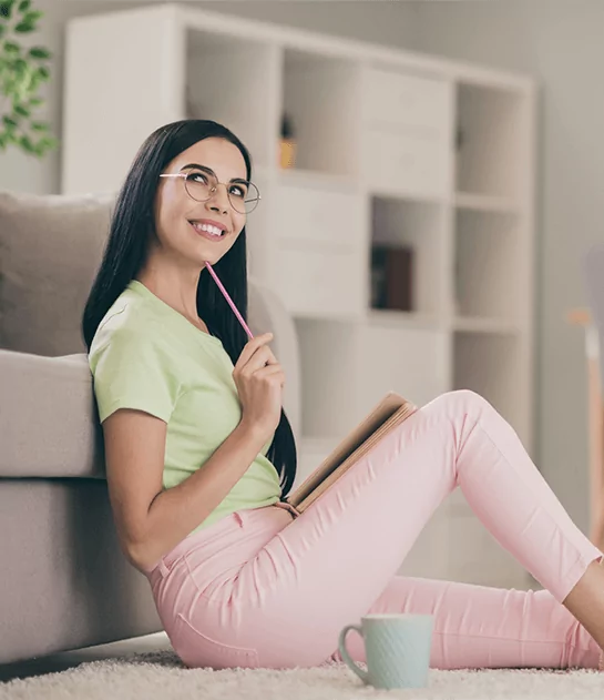 A smiling woman in a lime green shirt and pink pants sitting on the floor while brainstorming with a notebook, representing professional creative consultation and author development services.