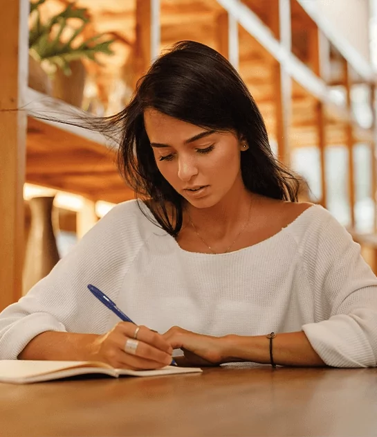 A woman with long dark hair writing in a notebook at a large wooden table.