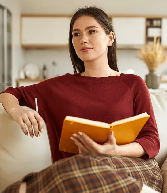 A woman in a burgundy sweater sitting on a couch and holding an open yellow notebook, illustrating professional author coaching and creative manuscript development.