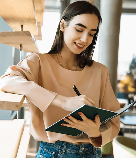 A smiling young woman in a beige top standing in a library and writing in a spiral notebook, representing on-site research and creative library services for authors.
