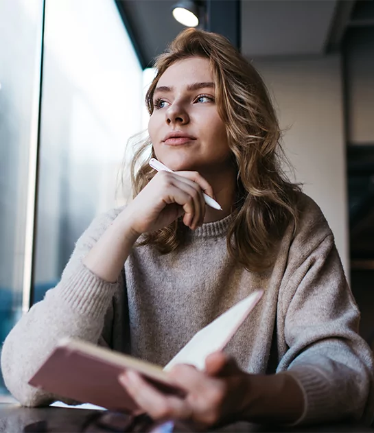A thoughtful woman in a beige sweater looking out a window while holding a pen and notebook, representing reflective writing and personalized author coaching services.