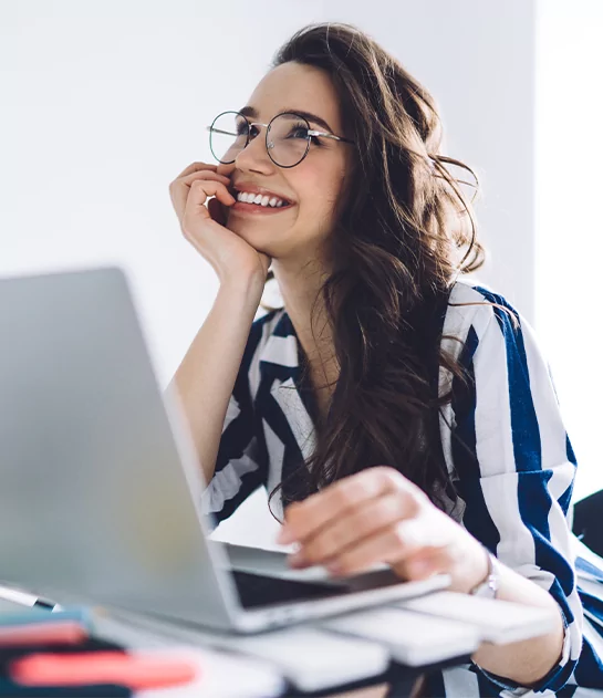 A smiling woman with glasses and a striped shirt looking up happily while working on her laptop