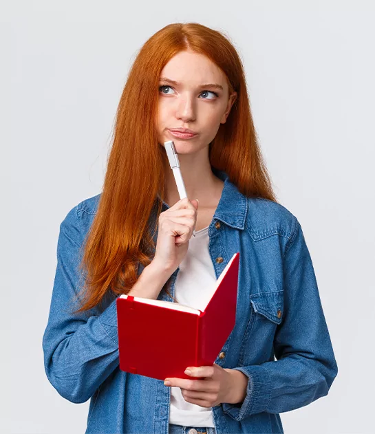 A young woman with long red hair and a denim shirt looking up thoughtfully while holding a pen to her chin and an open red notebook.