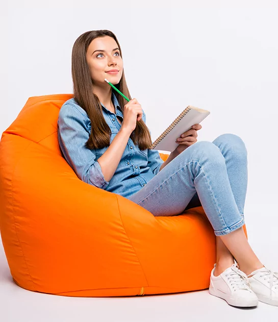 A thoughtful young woman sitting in an orange bean bag chair with a notebook and pencil, illustrating creative book development and author coaching.