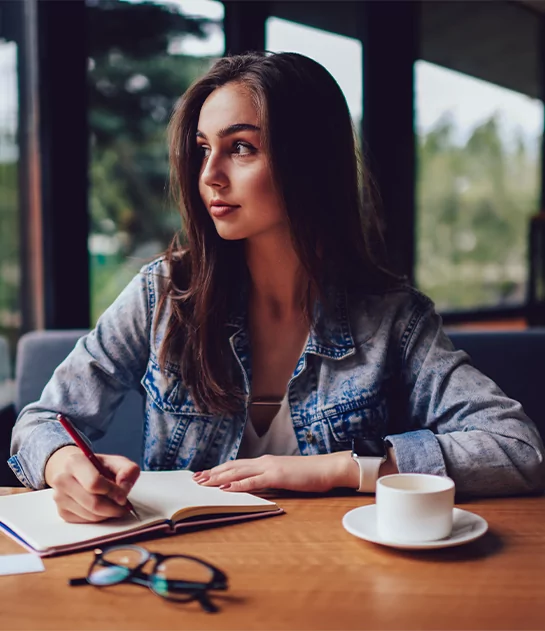 A young woman with long brown hair wearing a denim jacket, sitting at a wooden cafe table and writing in an open notebook while looking thoughtfully out a window.