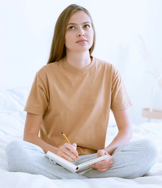 A young woman sitting cross-legged on a bed, looking upward thoughtfully while writing in a notebook, representing personalized creative coaching and author development services.