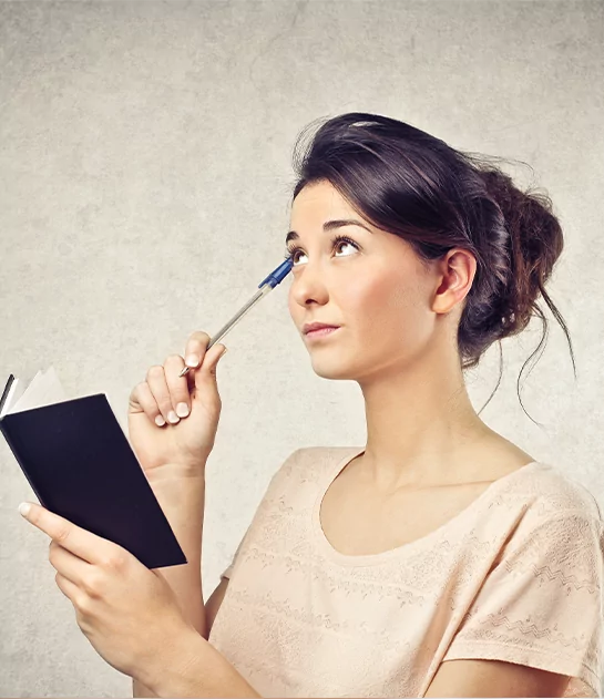 A woman with her hair in a bun looking up thoughtfully while holding a pen to her temple and carrying a small black notebook, symbolizing creative ideation and professional manuscript development services.