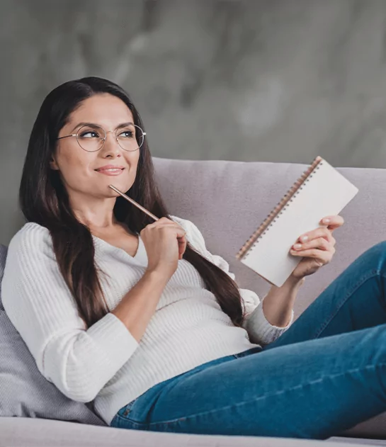 Thoughtful woman with glasses holding a spiral notebook while relaxing on a couch, representing professional author coaching and creative writing support.