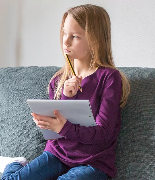 A young girl in a purple shirt sitting on a gray sofa, holding a pencil to her chin and looking thoughtfully into the distance while holding a drawing pad, representing personalized youth literature development and creative writing for children.