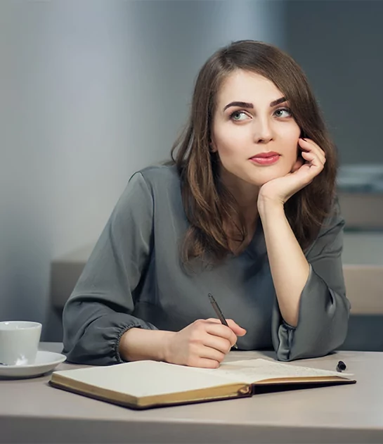 A thoughtful woman leaning on her hand while writing in an open journal at a table, symbolizing professional creative writing and storytelling services.