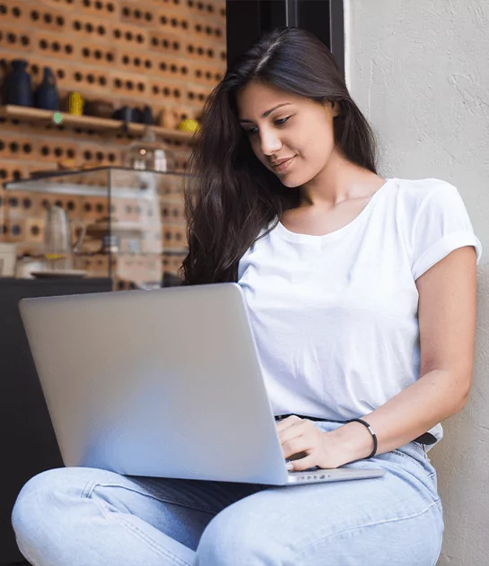 A young woman sitting cross-legged while smiling and working on a laptop against a wall.