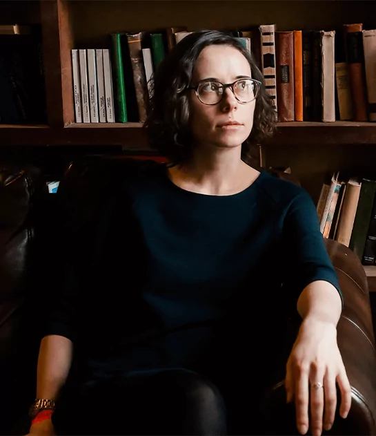 A woman with glasses sitting on a leather chair in front of a bookshelf, illustrating professional literary analysis and book curation services.