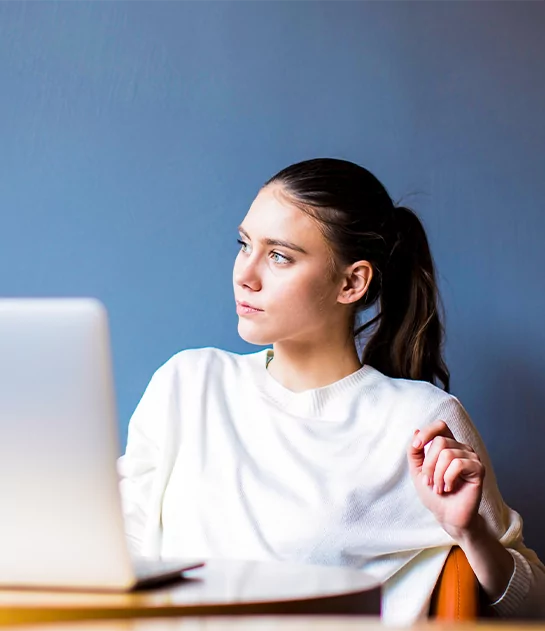 A young woman with a ponytail in a white sweater looking thoughtfully away from her laptop, representing professional freelance writing and digital content strategy.