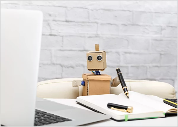 A small cardboard robot holding a fountain pen next to a laptop and notebook, symbolizing AI-assisted writing and modern publishing technology.