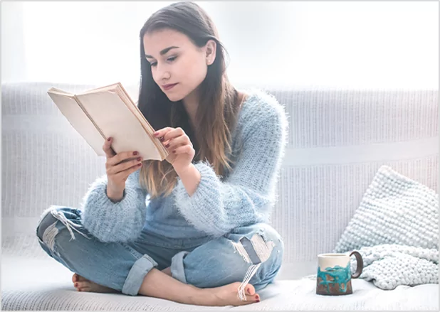 A woman in a fuzzy blue sweater sitting cross-legged on a couch while reading a book, illustrating comfortable home-based reading and literacy services.