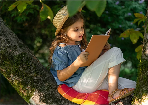 A young girl in a straw hat sitting in a tree while reading a book, representing children's book publishing and youth literacy programs.