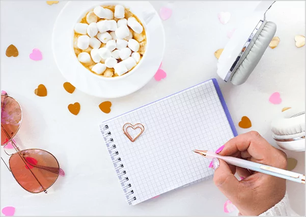 Top-down view of a hand writing in a grid notebook next to a cup of hot chocolate with marshmallows, symbolizing creative lifestyle blogging and personal brand storytelling.