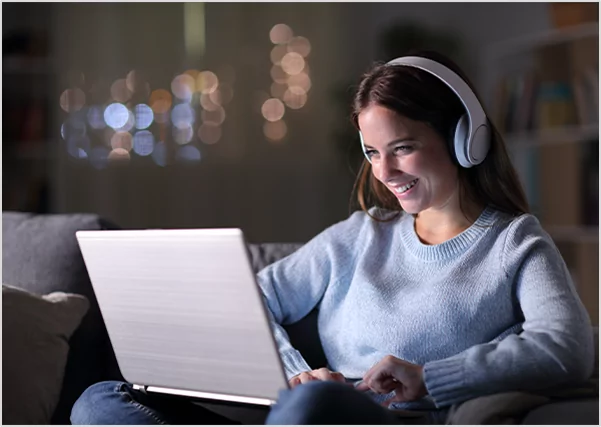 A smiling woman wearing white over-ear headphones, sitting on a couch at night while looking at a glowing laptop screen, representing professional audiobook production and digital narration services.
