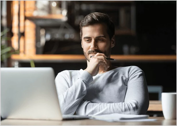 A thoughtful man with a beard sitting at a desk and looking at a laptop while holding a pen to his chin, representing professional developmental editing and manuscript critique services.