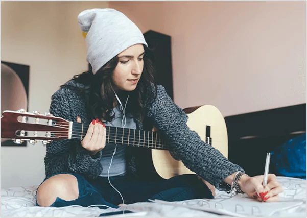 A young woman in a beanie and grey sweater sitting on a bed with an acoustic guitar, focused on writing music in a notebook.