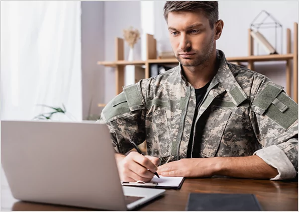 A man in a military camouflage uniform sitting at a desk, looking intently at a laptop screen while writing on a clipboard, symbolizing specialized military biography and memoir writing services.