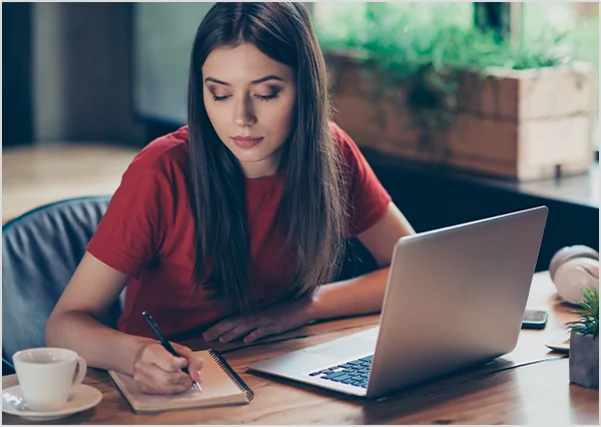 Focused woman in a red shirt writing in a notebook next to a laptop, illustrating professional manuscript development and writing services.