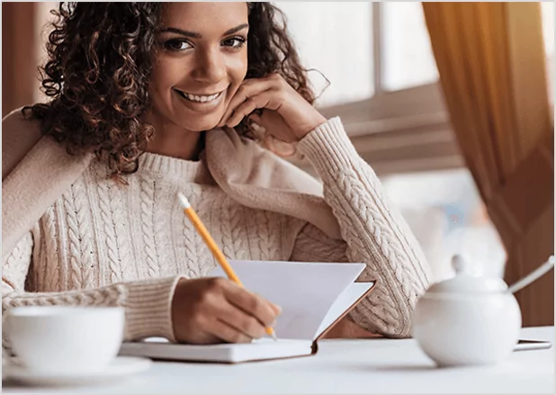 A smiling woman with curly hair wearing a cream cable-knit sweater, sitting in a cafe and writing in a journal with a pencil.