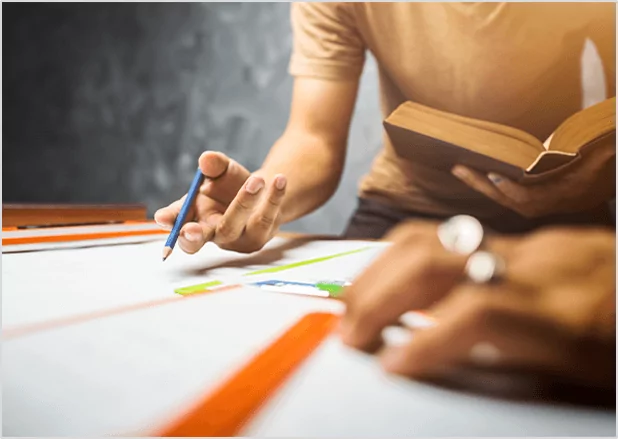 A person holding a blue pencil while reviewing documents and an open book, symbolizing professional editorial review and manuscript proofreading services.