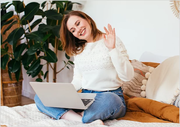 A smiling woman sitting cross-legged on a bed with a laptop, waving happily during a video call, representing personalized virtual author consultations and publishing support.