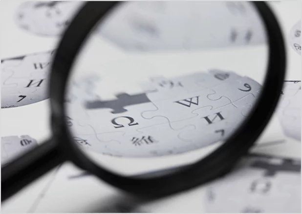 A magnifying glass held over pieces of a puzzle featuring the Wikipedia logo, symbolizing professional fact-checking and encyclopedic research services.