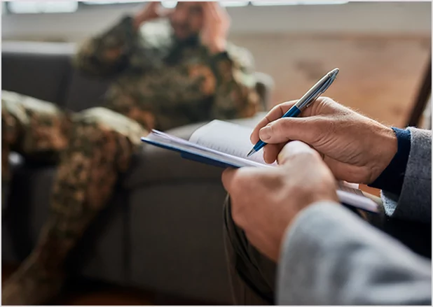 : A close-up of a professional interviewer's hands taking detailed notes in a journal while speaking with a veteran in the background, representing collaborative veteran memoir writing and interview-based biography services.