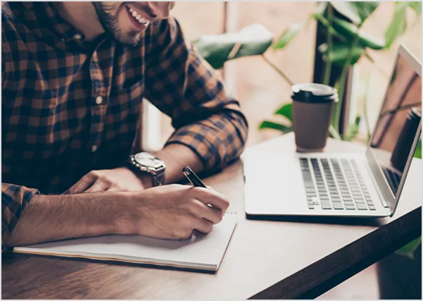 A smiling man in a plaid shirt writing in a notebook while working at a laptop, illustrating professional ghostwriting and publishing services.