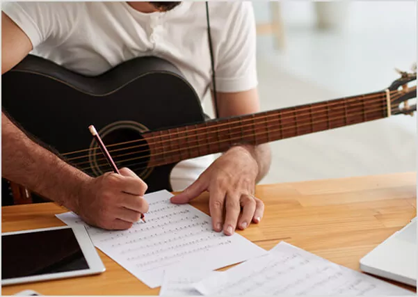 Close-up of a musician writing sheet music while holding an acoustic guitar, illustrating creative songwriting and publishing services.
