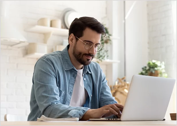 A man with glasses and a beard wearing a denim shirt, smiling while focused on typing on a laptop in a bright kitchen setting, representing professional remote author support and publishing services.