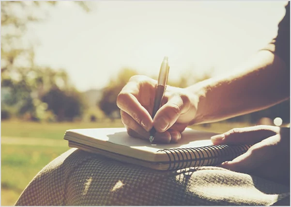 A close-up of a person's hand writing with a pen in a spiral notebook while sitting outdoors in a sunlit park.
