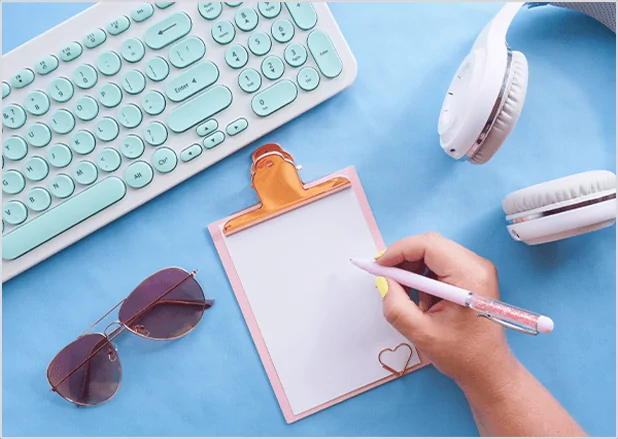A top-down view of a hand writing on a clipboard surrounded by a mint green keyboard, white headphones, and sunglasses, illustrating modern digital content creation and lifestyle blog publishing.