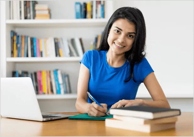 A smiling woman in a blue shirt writing at a desk with a laptop and bookshelves.