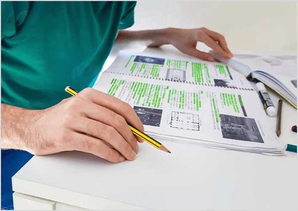 A woman with long dark hair writing in a notebook at a large, warm wooden table.