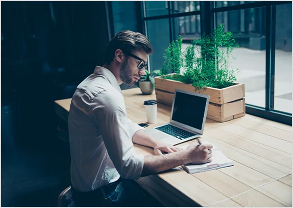 A man writing in a notebook while working at a laptop, illustrating professional writing and publishing.