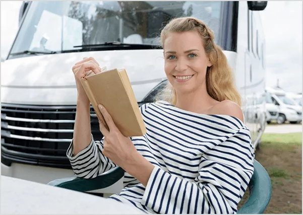 A smiling woman in a striped shirt sitting outside a camper van while reading a book, representing travel writing and nomadic lifestyle publishing services.